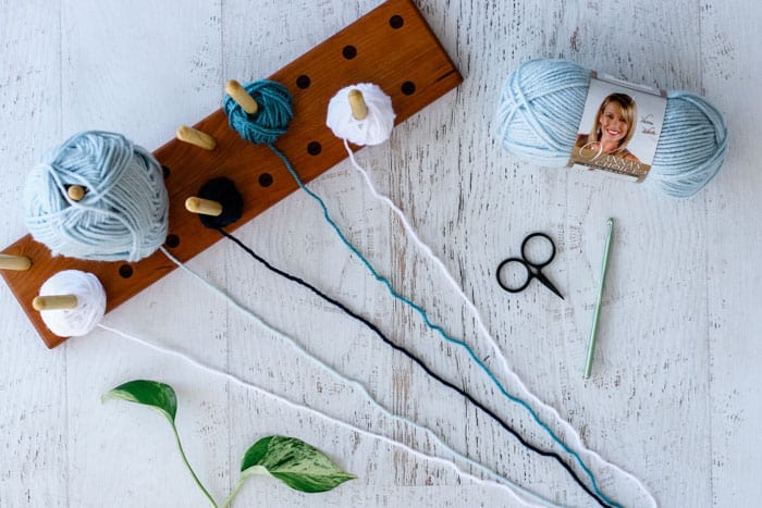 Flat lay of blue yarn scissors, a crochet hook, and a yarn wooden holder with various yarns and a leaf on a white background.
