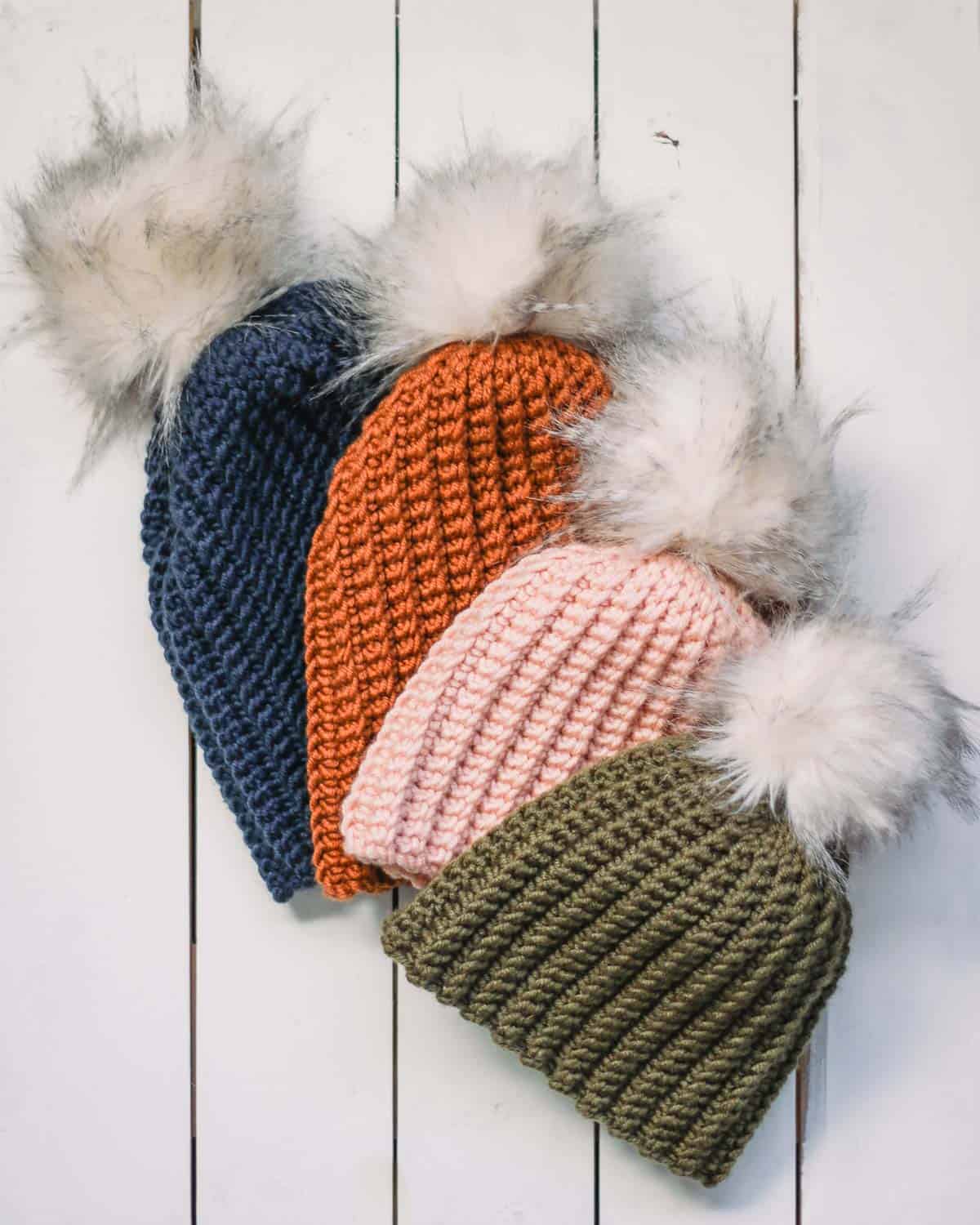 Four ribbed crochet beanies with fur pom poms laying on a white wood background. Hats in sizes baby-adult.
