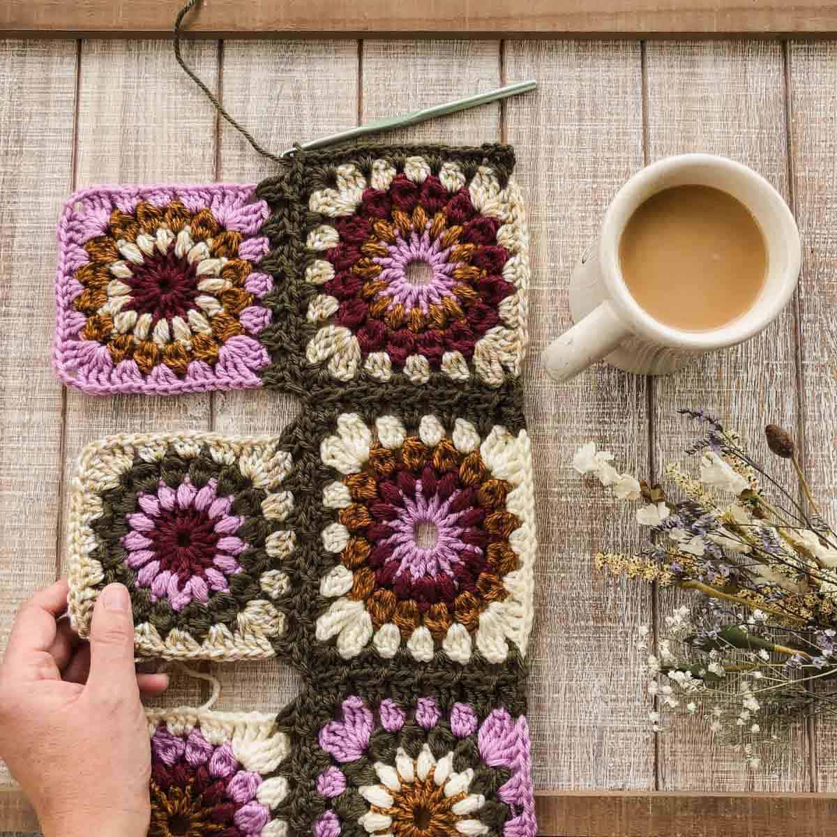 Flat lay of joined granny squares with a hand holding one square, a white mug with coffee, and dried flowers on the table.