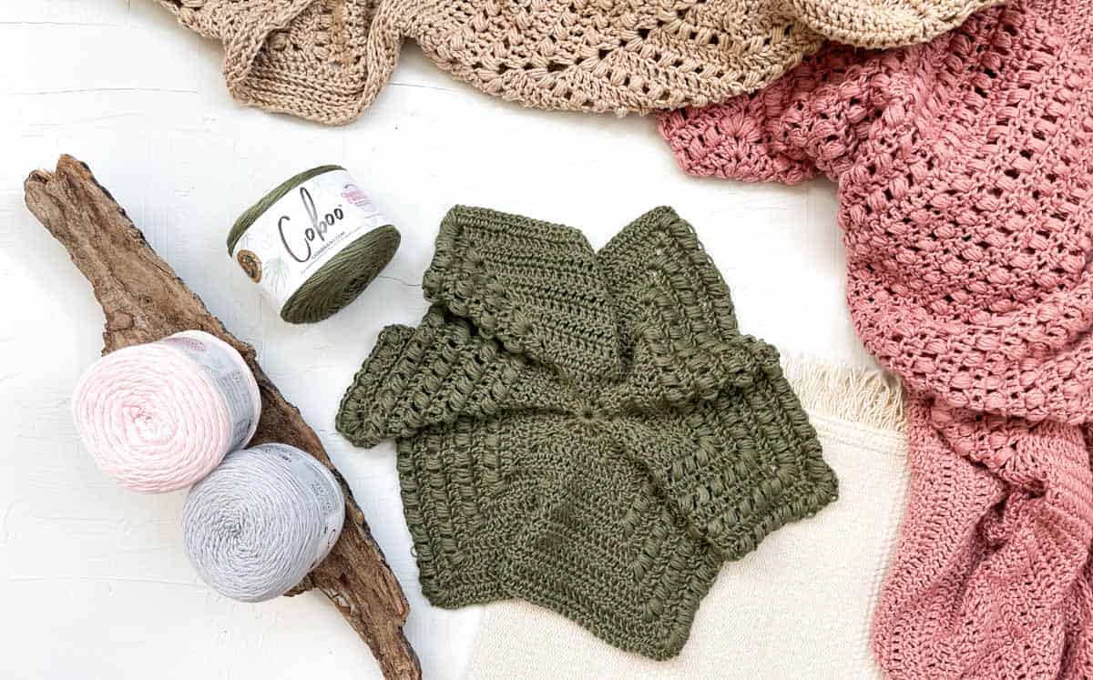 A WIP crochet hexagon sweater surrounded by a few balls of Lion Brand Coboo yarn and some sweaters that are also made from Coboo yarn.