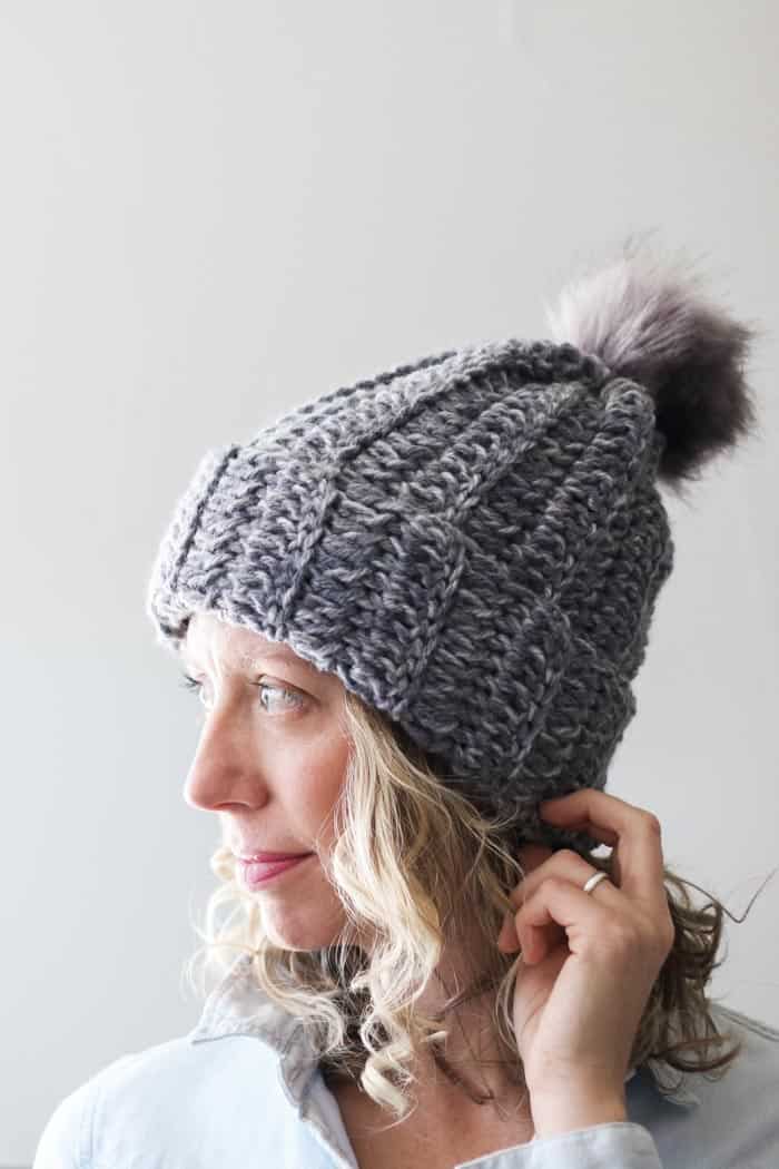 A woman looking in the side view while wearing a gray chunky crochet hat with a faux fur pom pom.