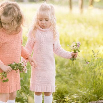Two girls standing in a field of green grass, wearing crochet sweater dresses and each holding a bouquet of wild flowers.