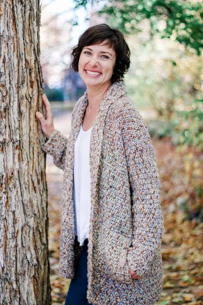 A woman smiling with her hands on a tree while wearing a crochet cardigan.